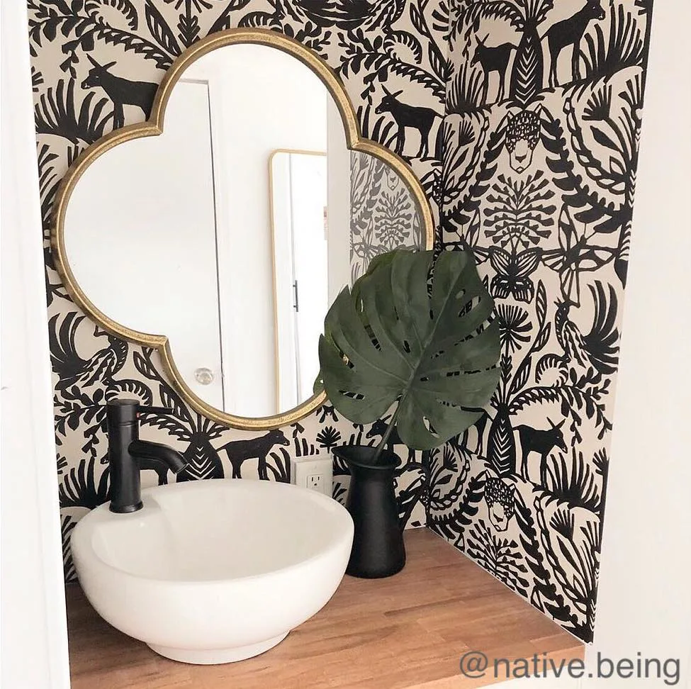 Decorative mirror hanging on a black and white wallpapered wall over a small bathroom sink.